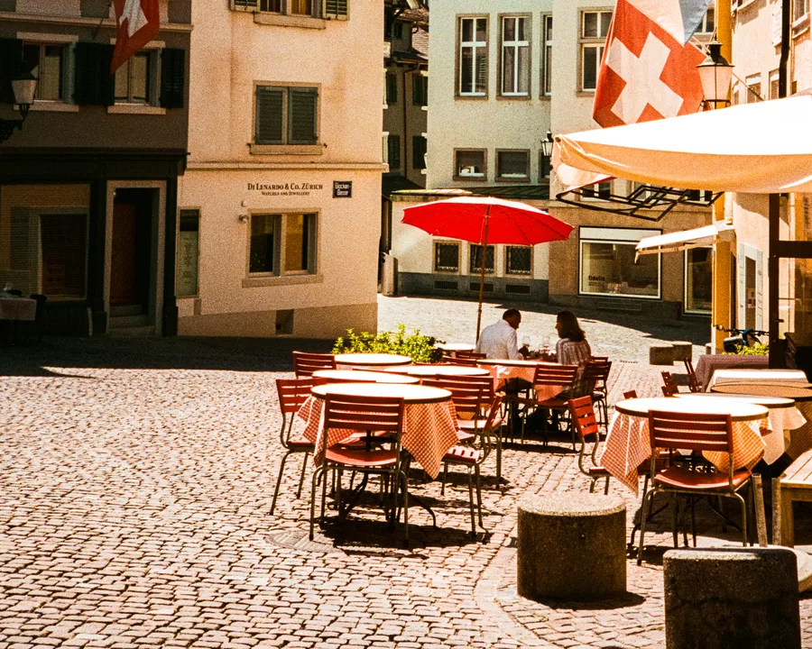 a couple at a café table under a red umbrella on a cobbled square, zürich