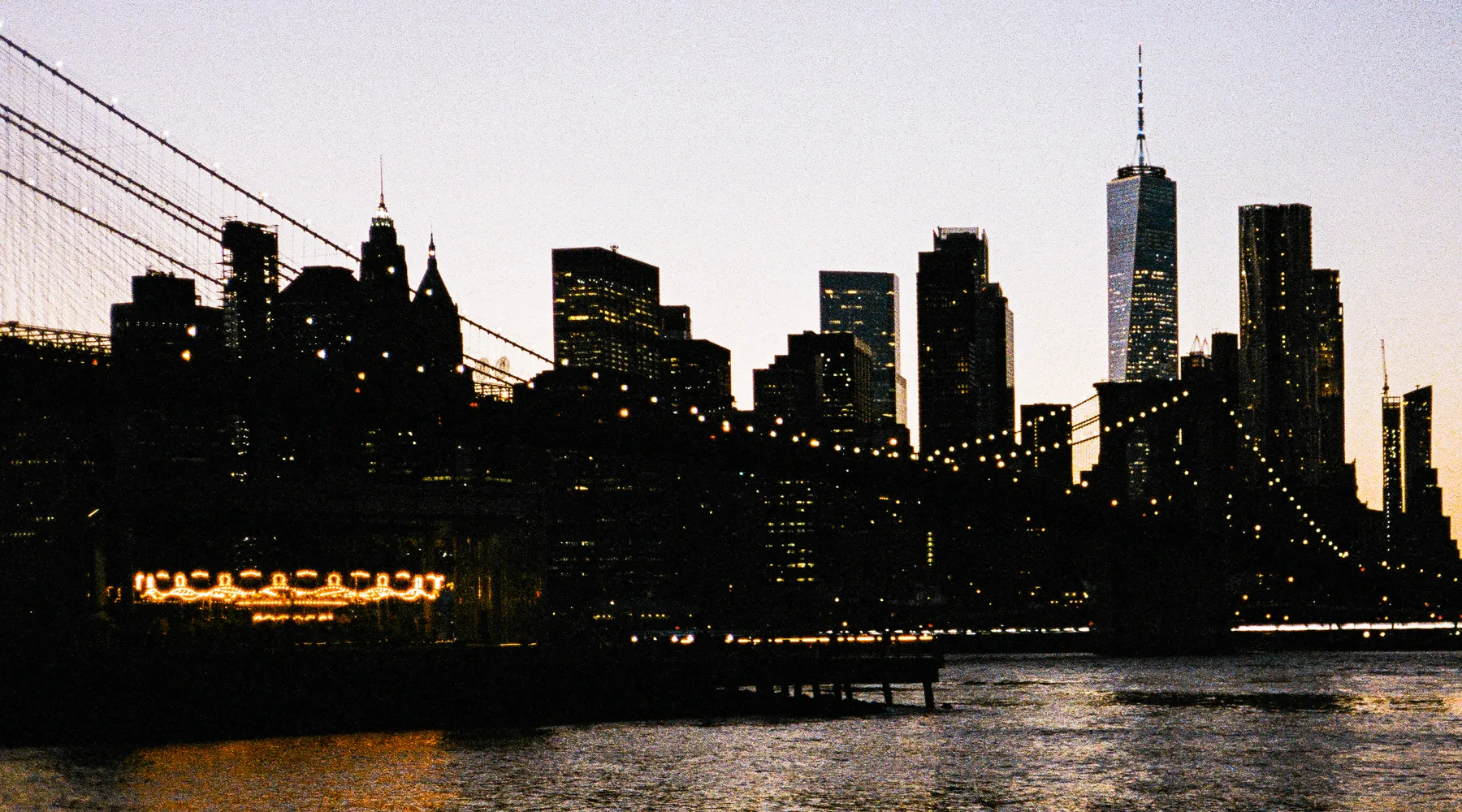 the brooklyn bridge and lower manhattan in silhouette at dusk, string lights glowing